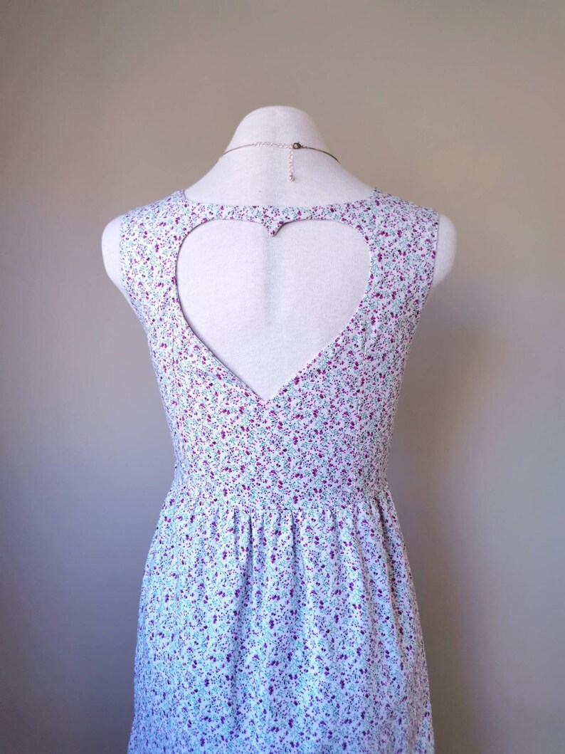 Peut inclure: Une robe blanche sans manches avec une d&eacute;coupe en forme de c&oelig;ur dans le dos. La robe a un motif floral dans les tons de violet, bleu et vert.