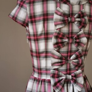 Puede incluir: Un vestido de cuadros blancos y rosas con tres grandes lazos en la espalda. Los lazos est&aacute;n hechos del mismo tejido de cuadros que el vestido.