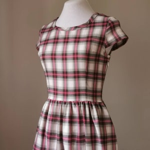 Puede incluir: Un vestido blanco con estampado de cuadros rosa y negros. El vestido tiene un escote redondo, mangas cortas y un corpi&ntilde;o ajustado. La falda est&aacute; fruncida en la cintura y llega hasta la rodilla.
