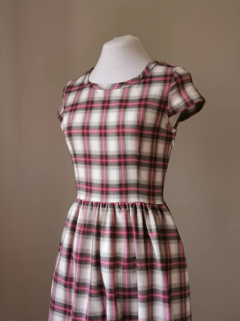 Puede incluir: Un vestido blanco con estampado de cuadros rosa y negros. El vestido tiene un escote redondo, mangas cortas y un corpi&ntilde;o ajustado. La falda est&aacute; fruncida en la cintura y llega hasta la rodilla.