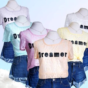 May include: Five lace tops with the text "Dreamer" in black lettering. The tops are in various pastel colors, including light blue, pink, yellow, and peach. Each top is paired with denim shorts.