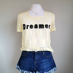 May include: A yellow lace top with short sleeves and the word "Dreamer" printed in black. The top is paired with blue denim shorts.