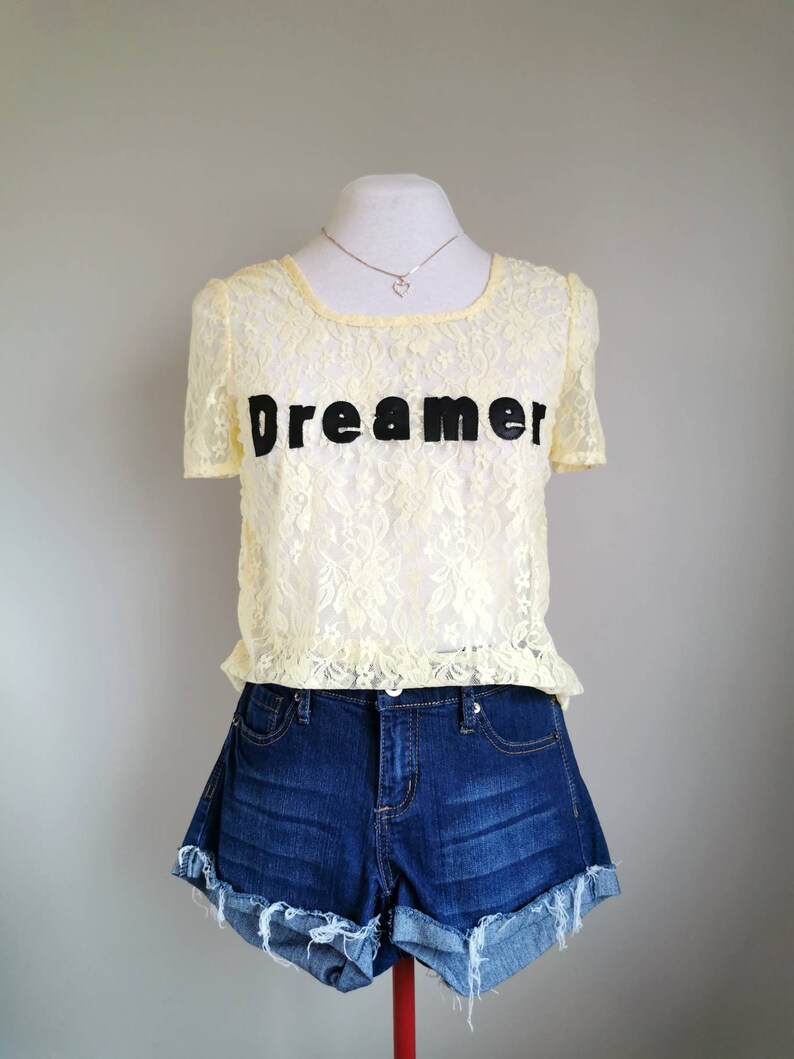 May include: A yellow lace top with short sleeves and the word "Dreamer" printed in black. The top is paired with blue denim shorts.