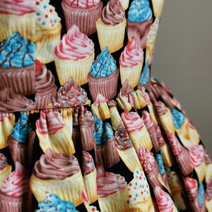 K&ouml;nnte beinhalten: Ein schwarzer Stoff mit einem sich wiederholenden Muster aus bunten Cupcakes. Die Cupcakes sind mit Streuseln, Zuckerguss und Kirschen verziert.