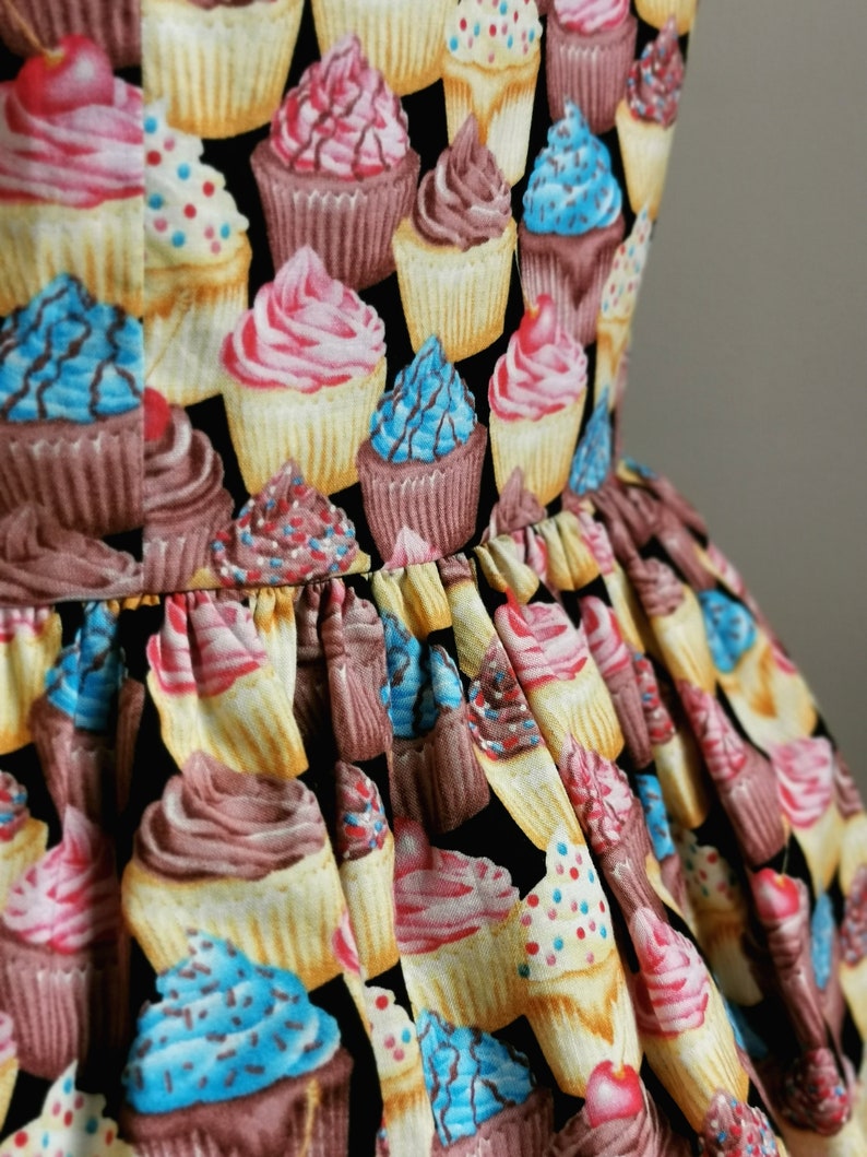 K&ouml;nnte beinhalten: Ein schwarzer Stoff mit einem sich wiederholenden Muster aus bunten Cupcakes. Die Cupcakes sind mit Streuseln, Zuckerguss und Kirschen verziert.