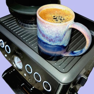 May include: A ceramic mug with a blue, purple, and white glazed exterior sits on a silver espresso machine. The mug is filled with coffee, and the machine has illuminated buttons and a pressure gauge.