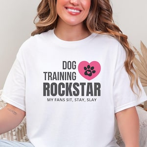 May include: White t-shirt with the text "DOG TRAINING ROCKSTAR" in black and a pink heart with a paw print. The text "MY FANS SIT, STAY, SLAY" is below the main text. The shirt is worn by a person.