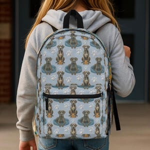 May include: A light blue backpack featuring a repeating pattern of dogs, dog beds, and paw prints. The backpack has a black zipper and straps. The person is wearing a gray hoodie.