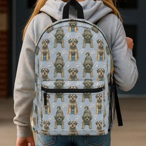 Könnte beinhalten: Hellblauer Rucksack mit einem sich wiederholenden Muster aus Cartoon-Hunden und Pfotenabdrücken. Der Rucksack hat einen schwarzen Griff, eine schwarze Netztasche an der Seite und eine Vordertasche mit schwarzem Reißverschluss. Die Person trägt einen grauen Kapuzenpullover.
