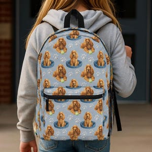 Könnte beinhalten: Hellblauer Rucksack mit sich wiederholendem Muster aus braunen Cocker Spaniels in blauen Hundebetten, Tennisbällen und weißen Pfotenabdrücken. Er hat schwarze Träger und eine Vordertasche.