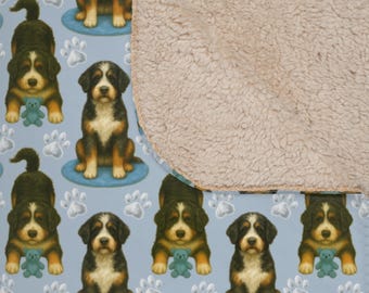 May include: A reversible blanket featuring a pattern of Bernese Mountain Dog puppies with blue teddy bears and paw prints on a light blue background. The reverse side is a soft, tan sherpa material.