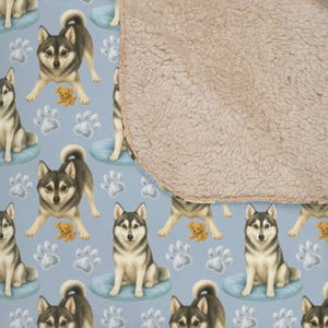 May include: A light blue blanket featuring a repeating pattern of watercolor-style Siberian huskies, small tan puppies, and paw prints. The blanket has a soft, tan sherpa fleece backing.