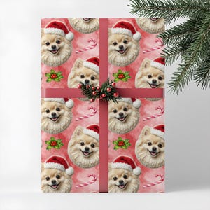 May include: Christmas-themed gift wrap with a repeating pattern of Pomeranian dogs in Santa hats, candy canes, and holly on a pink watercolour background. The gift is wrapped with a red ribbon and a sprig of berries.
