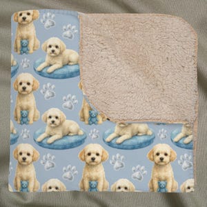 May include: A light blue blanket with a repeating pattern of cream-coloured puppies, blue teddy bears, and paw prints. The blanket has a soft, plush, tan sherpa lining on the reverse side. The puppies are sitting and lying on blue cushions.