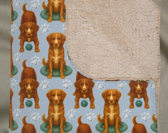 Könnte beinhalten: Hellblaue Decke mit einem sich wiederholenden Muster aus braunen Hunden auf grünen Kissen, grünen Bällen und weißen Pfotenabdrücken. Die Rückseite ist ein beiges, plüschiges Material.
