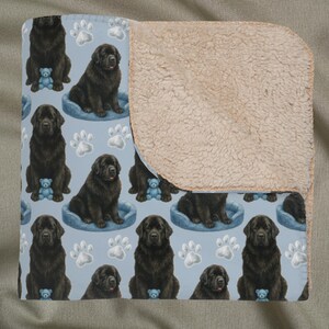 Peut inclure: Une couverture avec un motif répété de chiens Terre-Neuve noirs, de lits bleus pour chiens, d'empreintes de pattes et d'ours en peluche bleus sur fond bleu clair. Le verso est en sherpa beige doux.