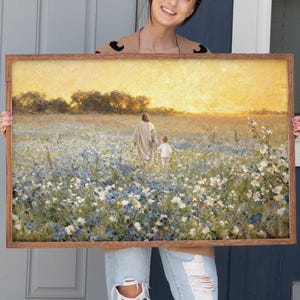 Puede incluir: Una impresión artística enmarcada que representa a una persona con una túnica blanca tomados de la mano con un niño, caminando por un campo de flores azules y blancas. La obra de arte tiene una cálida puesta de sol dorada en el fondo y un marco de madera marrón.