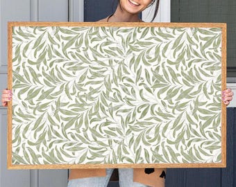 May include: A rectangular framed print featuring a repeating pattern of green leaves on a white background. The frame is made of light brown wood. The print is held by a person.