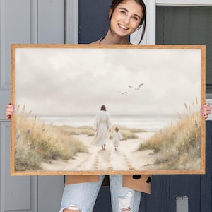 Puede incluir: Una impresión artística enmarcada representa una escena de playa serena. Dos figuras, un adulto y un niño, caminan de la mano por un sendero arenoso hacia el océano. La obra de arte presenta tonos suaves y apagados y un marco de madera.