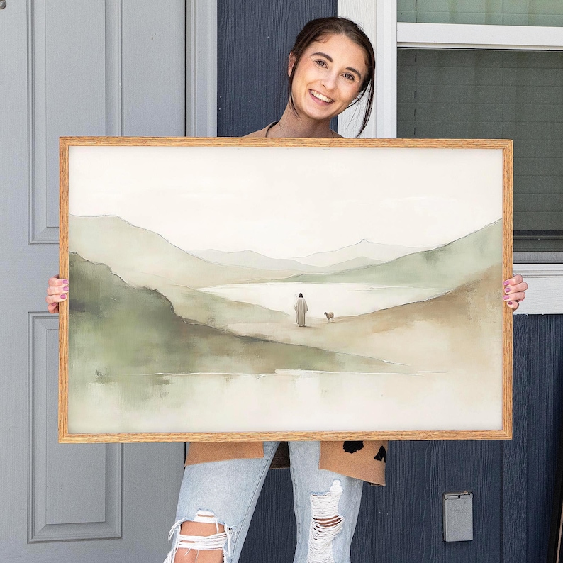 May include: A framed watercolor landscape painting with a light wood frame. The artwork depicts a figure with a dog walking towards a body of water, with mountains in the background. The color palette is soft, featuring muted greens, browns, and whites.