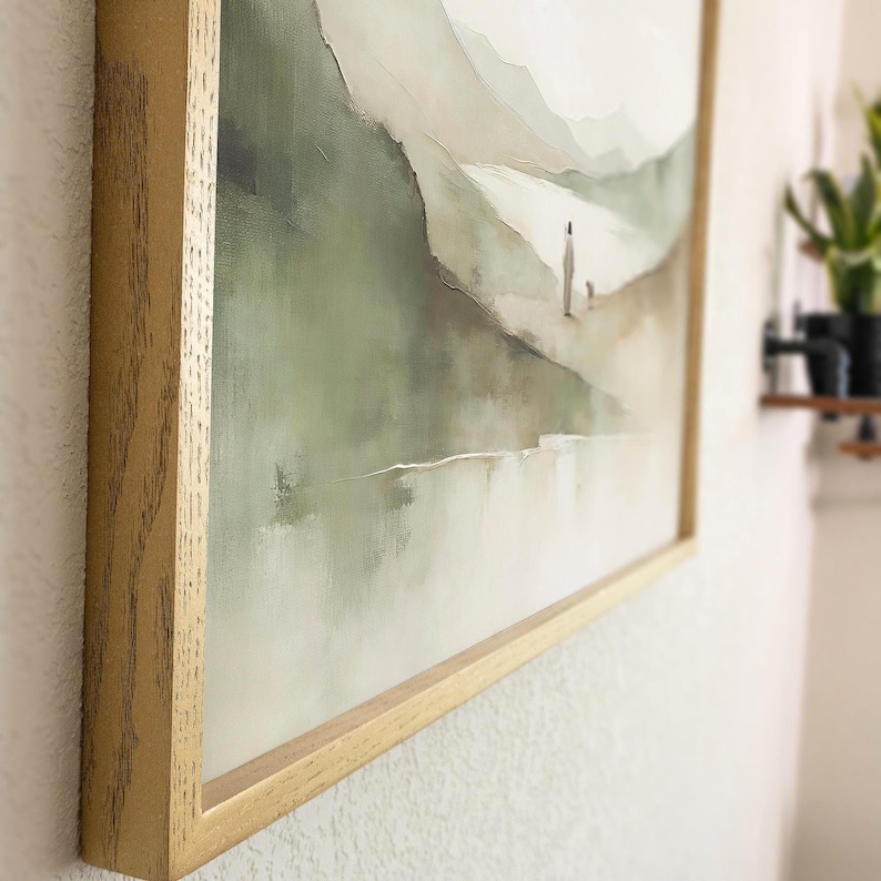 May include: A framed landscape painting with a natural color palette. The artwork depicts a serene scene with rolling hills, a body of water, and a figure. The frame is a light wood color, adding a touch of warmth to the piece. The painting is mounted on a white wall.