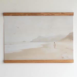 Könnte beinhalten: Ein Kunstdruck einer Strandszene, befestigt mit magnetischen Holzleisten. Das Kunstwerk zeigt eine Person, die an einem Sandstrand auf das Meer zugeht, mit Bergen im Hintergrund und Vögeln am Himmel. Die Farbpalette ist weich und gedämpft, mit Beige- und Grautönen.