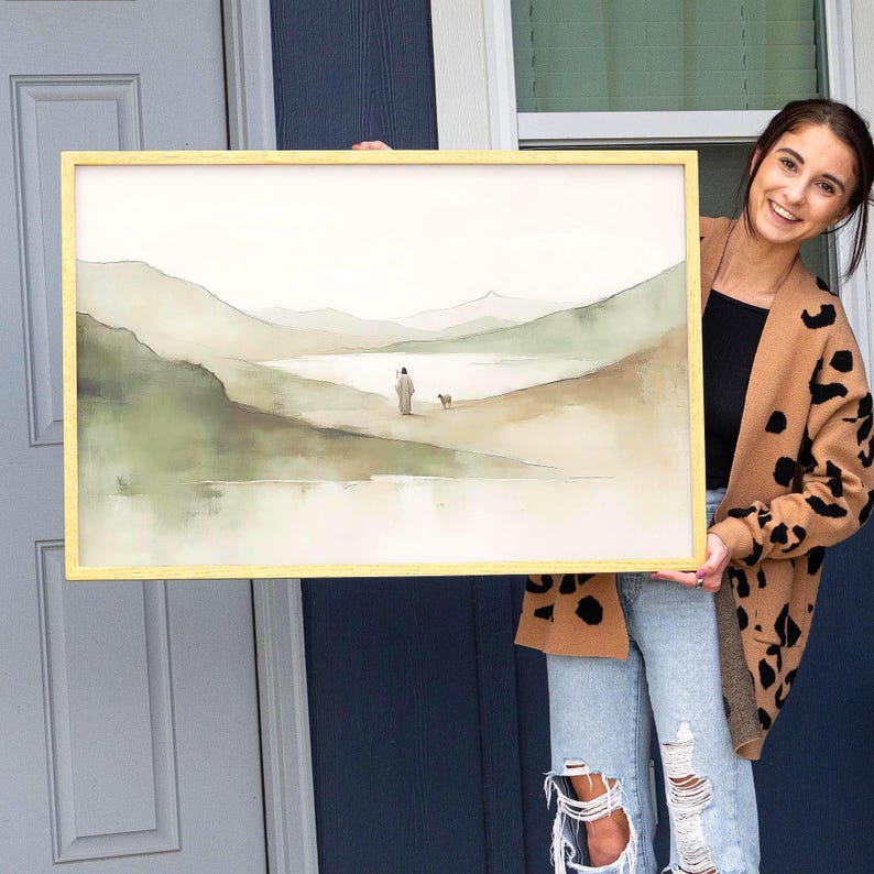May include: Watercolor landscape art print in a gold frame. The artwork depicts a figure with a dog walking near a body of water, with rolling hills in the background. The color palette includes shades of green, beige, and gray.