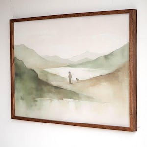 May include: Watercolor landscape art print in a brown wooden frame. The artwork depicts a person in a long robe with a small dog, walking along a path near a lake, with green mountains in the background. The color palette is soft, with muted greens, browns, and beige.