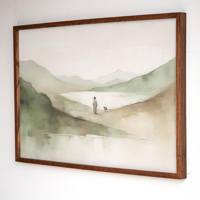 May include: Watercolor landscape art print in a brown wooden frame. The artwork depicts a person in a long robe with a small dog, walking along a path near a lake, with green mountains in the background. The color palette is soft, with muted greens, browns, and beige.