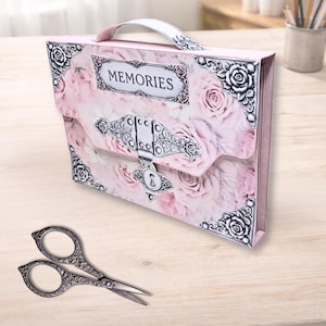 May include: A pink scrapbook with a floral rose design and the word "MEMORIES" on the front. The scrapbook has a silver clasp and handle. A pair of decorative scissors with floral detailing is in the foreground.