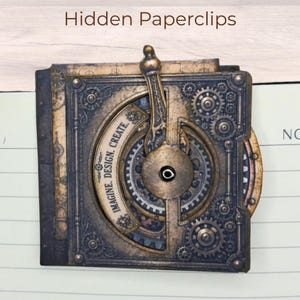 Può includere: Un portaclip in stile steampunk con intricati disegni di ingranaggi e ruote dentate nei toni del bronzo e dell'oro. Il testo "IMAGINE. DESIGN. CREATE." è visibile. Le parole "Hidden Paperclips" sono in alto.