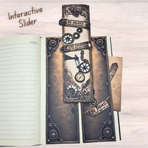 May include: An interactive slider bookmark featuring a brown and black mechanical gear design with clock motifs. Text includes BE BRAVE WHEN YOU ARE SCARED, STAY FOCUSED ON YOUR GOALS, and BE BADASS EVERY SINGLE DAY. The bookmark is placed on a lined notebook.