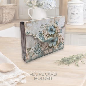 Puede incluir: Un tarjetero de recetas con un diseño floral desgastado en tonos azules, crema y verdes. El soporte tiene una solapa curva y una pequeña etiqueta metálica que dice "RECIPES". El texto "RECIPE CARD HOLDER" está en la parte inferior.