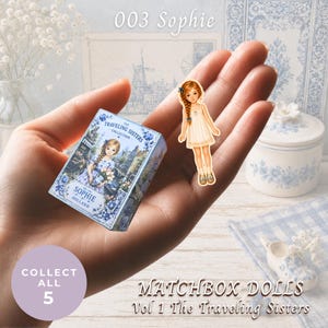 May include: A small matchbox labeled 003 Sophie The Traveling Sisters Collection with blue floral patterns and a matching paper doll figure with braided hair wearing a cream dress. The text reads 003 Sophie, Collect All 5, and Matchbox Dolls Vol 1 The Traveling Sisters.