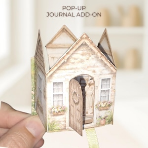 Puede incluir: Un complemento de diario emergente que presenta una casa en miniatura con una puerta abierta y detalles florales. La casa tiene una apariencia envejecida detallada con un tejado a dos aguas y jardineras. El texto "POP-UP JOURNAL ADD-ON" está impreso en la parte superior.