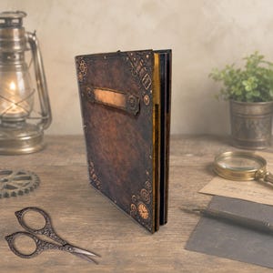 May include: A rectangular dark brown journal with a textured cover featuring metallic gear and clockwork embellishments at the corners and a central rectangular plaque. The spine is black, and the edges of the pages are finished in a metallic gold tone.