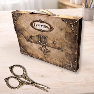 Pode incluir: Uma caixa de armazenamento em estilo vintage com a palavra "Ephemera" na frente, com detalhes de canto de metal ornamentados e um fecho. Um par de tesouras decorativas com detalhes florais está em frente à caixa.
