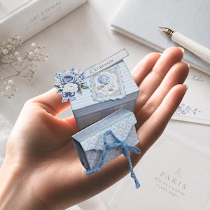 Può includere: Una piccola scatola a righe blu e bianche con il testo L'ÉCRIVAIN, decorata con un bouquet floreale e un'illustrazione di ortensia, abbinata a una busta più piccola coordinata con un laccio blu e un motivo geometrico.
