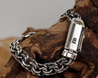 May include: A silver bracelet with a chunky chain link design. The bracelet has a secure clasp and is displayed on a piece of wood. The metal has a polished finish, reflecting light.