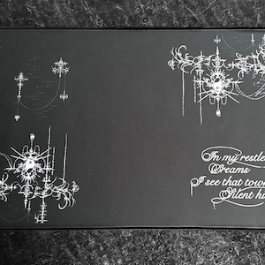 May include: A rectangular black desk mat featuring intricate white line art designs on the left and right sides. The right side includes the white text: In my restless dreams I see that town Silent hill. The design features ornate, spindly patterns.