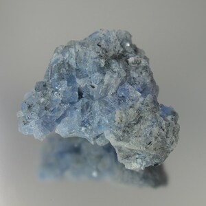 May include: A close-up of a light blue mineral specimen with a crystalline structure. The mineral has a rough, textured surface with areas of darker gray and black inclusions. The overall shape is irregular and jagged, with a reflective quality.