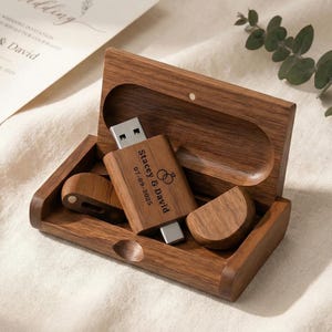 May include: A rectangular wooden box with a hinged lid containing a wooden USB flash drive. The drive is engraved with the text Stacey & David and the date 07-09-2025, accompanied by a small icon of two interlocking wedding rings.