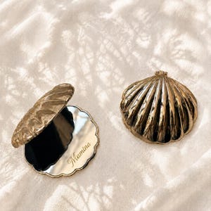 May include: Two gold-toned shell-shaped compact mirrors. One is closed, showing a ribbed exterior. The other is open, revealing a circular mirror with the name Mamina engraved in cursive script on the reflective surface and a hinged lid.