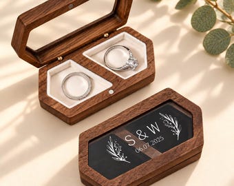 May include: Two hexagonal wooden ring boxes with dark brown wood grain. One box is open, revealing two white velvet-lined compartments holding silver rings. The other box is closed, featuring a glass lid with white text S & W and 06.07.2025.