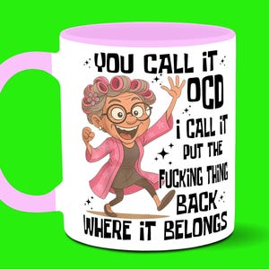May include: White ceramic mug with a pink handle and rim, featuring a cartoon of an elderly woman with the text "YOU CALL IT OCD I CALL IT PUT THE FUCKING THING BACK WHERE IT BELONGS".