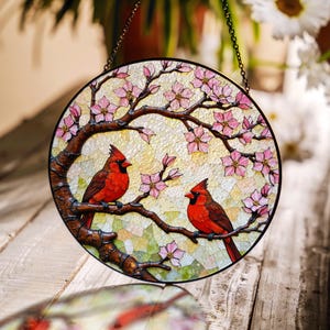 May include: A stained glass suncatcher featuring two red cardinals perched on a branch with pink blossoms. The suncatcher is round and has a chain for hanging.