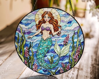 Puede incluir: Una ventana de vidrio emplomado colgante presenta una sirena con cabello largo rojo, un sujetador de concha de mar púrpura y una cola verde y azul. La sirena está rodeada de olas azules y moradas y algas verdes.
