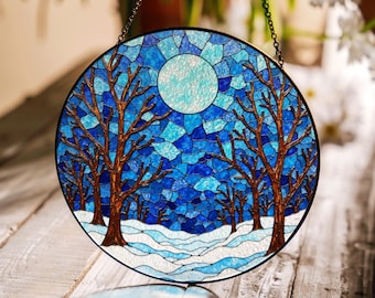 Op de afbeelding: Rond gebrandschilderd glaspaneel met een winterlandschap. Het ontwerp toont bruine bomen zonder bladeren, een volle maan en een besneeuwd landschap, alles tegen een mozaïek van blauw en turkoois glas. Een zwarte ketting is bevestigd om op te hangen.