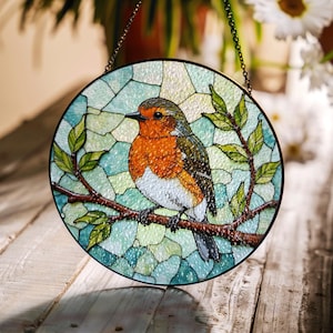 Op de afbeelding: Een hangend glas-in-loodraam toont een roodborstje dat op een tak zit met groene bladeren. Het roodborstje is afgebeeld in tinten oranje, rood, wit en bruin. De achtergrond is een mozaïek van blauw, groen en wit glas.