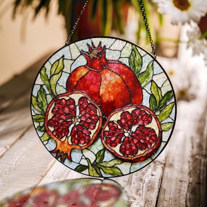 May include: A stained glass window hanging with a round design featuring a red pomegranate with green leaves. The pomegranate is cut open to reveal the red seeds inside.